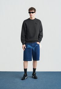 Νέος άνδρας που φοράει μαύρο φούτερ Puma, μπλε τζιν σορτς Puma, μαύρες κάλτσες και αθλητικά παπούτσια, καθώς και ορθογώνια μαύρα γυαλιά ηλίου, στέκεται σε μπλε πάτωμα.