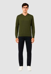 Maglione verde a scollo a v con polsini e orlo a coste, abbinato a pantaloni neri e sneakers bianche. Il modello si trova davanti a uno sfondo grigio neutro.