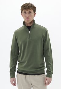 Pullover verde a mezza zip realizzato in tessuto liscio, con maniche lunghe e vestibilità rilassata, indossato sopra una maglietta nera.