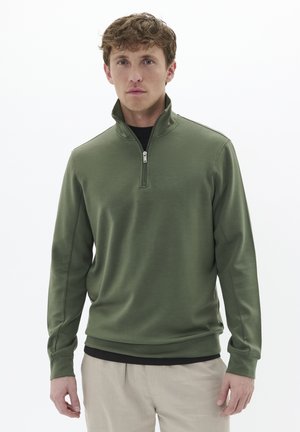 Pullover verde a mezza zip realizzato in tessuto liscio, con maniche lunghe e vestibilità rilassata, indossato sopra una maglietta nera.