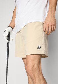 Beige shorts laget av lett materiale, med en brodert svart logo, sidelommer og en avslappet passform. Golfkølle i hånden.