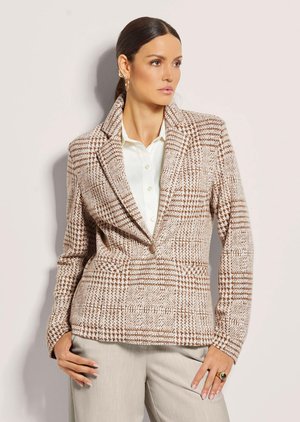 Vrouw met donker haar in een paardenstaart, die een beige en bruine geruite blazer draagt over een wit overhemd met beige broek, poseert tegen een lichte muur.