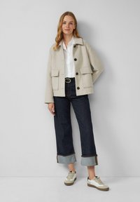 Beige Wolljacke aus einem Wollgemisch mit zwei großen Fronttaschen, Knopfverschluss und Kragen. Trägt man über einem weißen Hemd und kombiniert mit dunklen Jeans.