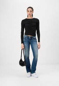 Svart langermet topp med rynkete frontdetalj, kombinert med mid-blue slitte jeans og hvite sneakers, holder en liten svart håndveske.
