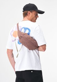 Braune Nylon-Schultertasche mit geschwungener Form, subtiler Textur und runden Akzenten, über den Körper getragen über einem weißen T-Shirt mit buntem floralen Grafikdesign.
