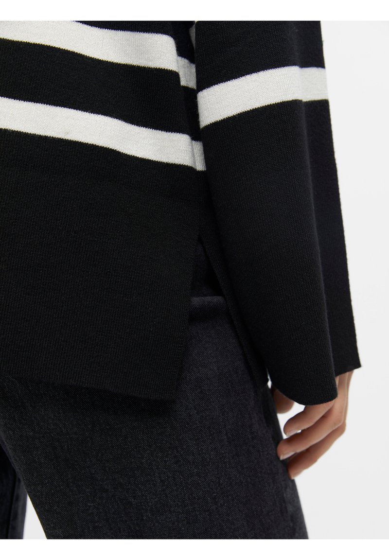 Pull rayé noir et blanc avec une coupe décontractée, texture en tricot, fentes sur les côtés et manches longues, associé à un pantalon sombre.