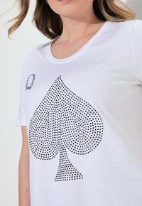 Camiseta blanca de algodón, con un gran diseño en forma de trébol negro creado con pequeños puntos y un símbolo de trébol adicional a la izquierda.