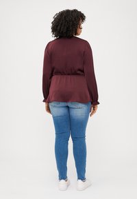 A person with curly hair, wearing a burgundy top and blue jeans, stands with their back to the camera against a white background.