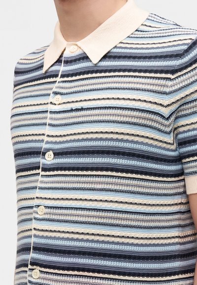 Homme portant une chemise à manches courtes boutonnée avec des rayures horizontales dans des nuances de bleu, noir, crème et blanc, avec un col crème.