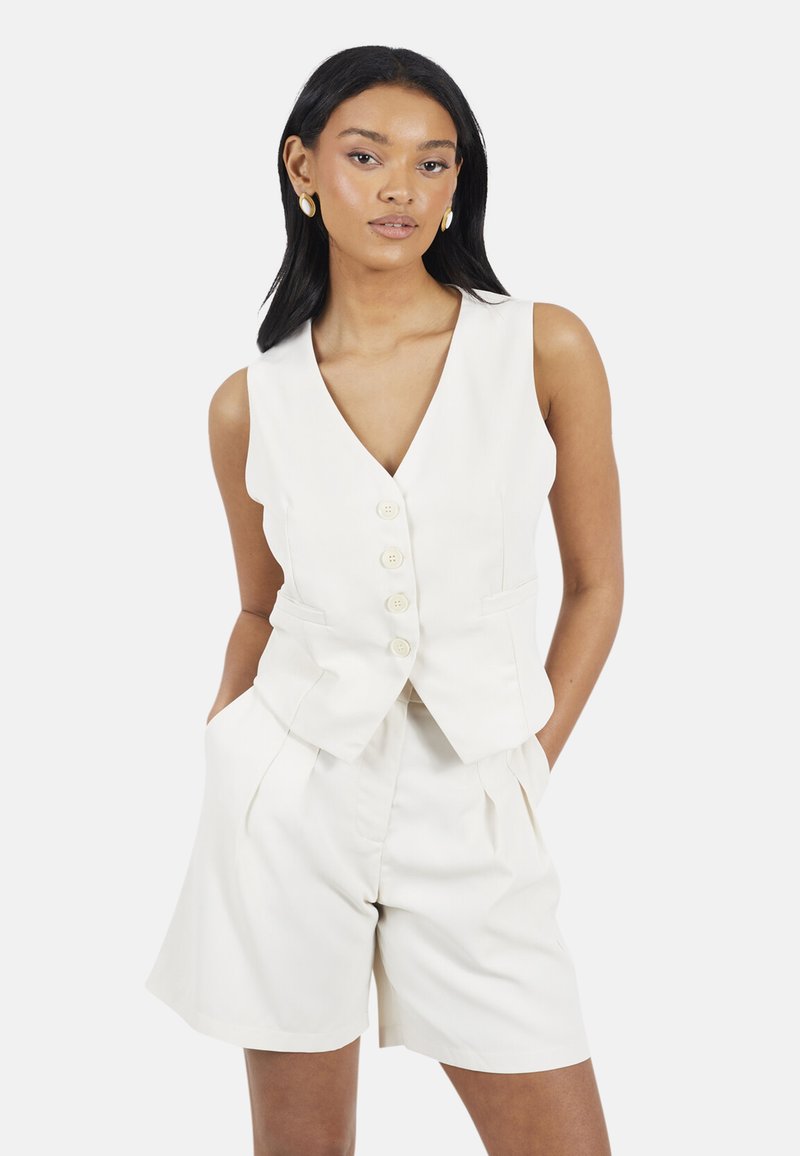 Vrouw met zwart haar, gekleed in een mouwloos wit vest met knopen en bijpassende hoge shorts, staand met handen in de zakken, neutrale achtergrond.