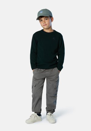 Sudadera verde, pantalones cargo gris con bolsillos laterales y zapatillas de color claro. También lleva una gorra gris con un logo circular.