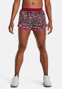 En atletisk person klädd i mönstrade maroon- och svarta shorts med vita och rosa accenter, står med benen isär mot en vit bakgrund.