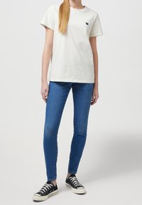 Camiseta blanca de algodón de manga corta con un logo azul marino en el pecho, combinada con jeans ajustados azules y zapatillas negras.
