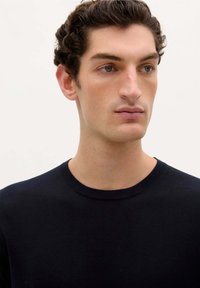 Marineblå crew-neck t-shirt lavet af blødt bomuld, med en glat tekstur og en ren halsudskæring, båret af en model med kort krøllet hår.