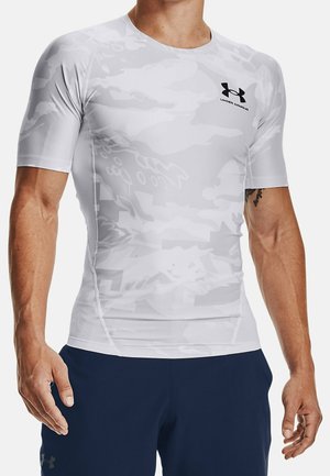 Mannelijke torso gekleed in een witte camouflerende, korte mouw Under Armour compressieshirt en donkerblauwe sportshorts, staand tegen een effen achtergrond.