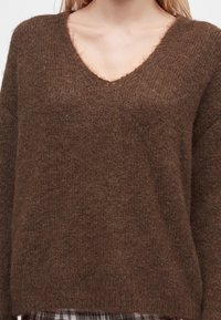 Femme portant un pull en maille doux marron à col en V et un pantalon à carreaux, montrant le haut du torse et l'encolure.