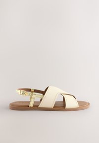 Sandale en cuir beige avec design à brides croisées, boucle dorée réglable et semelle plate marron. Texture lisse avec peu de détails.