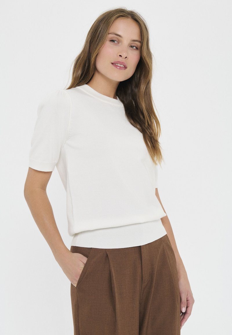 Cream-colored short-sleeve sweater with a fitted design and ribbed hem. Worn with brown tailored trousers, featuring a relaxed fit and side pockets.
