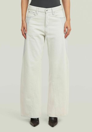 Personne portant un jean blanc taille haute à jambes larges, associé à des bottines noires à bout pointu et un haut noir, debout devant un fond uni.