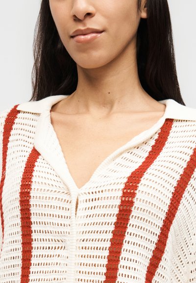 Γυναίκα που φοράει λευκό ανοιχτό πλεκτό ζακέτα με κάθετες κόκκινες ρίγες και γιακά, με ορατό το ντεκολτέ και μέρος του προσώπου και των μαλλιών της.