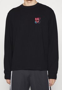Person, der ein schwarzes Langarmshirt mit einem bunten "HUGO"-Logo auf der oberen linken Brust trägt, kombiniert mit dunklen Hosen mit weißen Seitenstreifen.