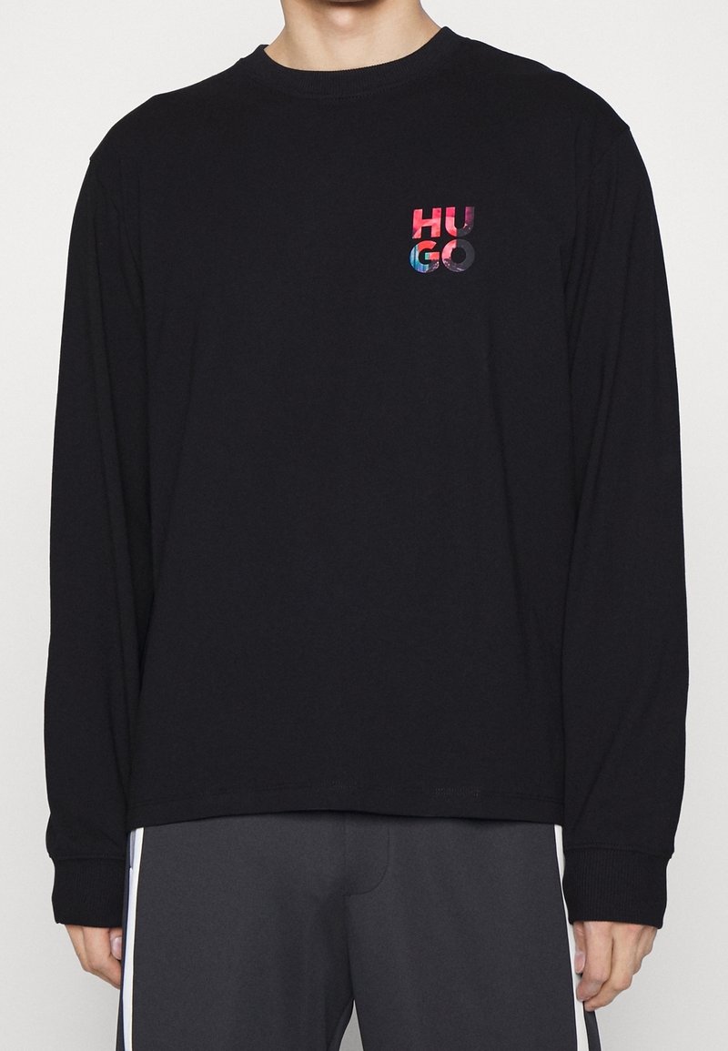 Person, der ein schwarzes Langarmshirt mit einem bunten "HUGO"-Logo auf der oberen linken Brust trägt, kombiniert mit dunklen Hosen mit weißen Seitenstreifen.