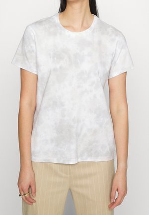 Kobieta w krótkim, jasnoszarym, malowanym metodą tie-dye t-shircie i beżowych spodniach w cienkie paski, stoi z rękami luźno opuszczonymi wzdłuż ciała.