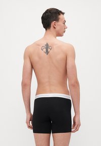 Boxeri negri din bumbac cu un elastic alb inscripționat "Copenhagen Studios." Prezintă un tatuaj decorativ pe partea superioară a spatelui.
