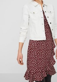 Kvinna som bär en vit knappad denimjacka över en vinröd klänning med vita oregelbundna prickar och volangkant.