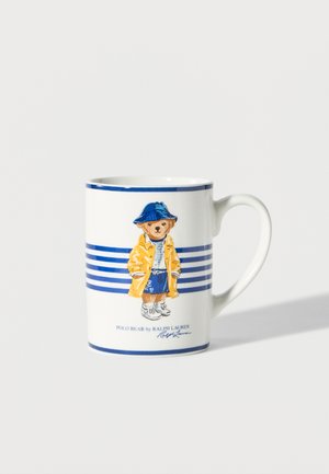 Biały ceramiczny kubek z niebieskimi paskami oraz ilustracją niedźwiedzia w żółtym płaszczu przeciwdeszczowym, niebieskich szortach, kapeluszu i tenisówkach, z napisem Polo Bear by Ralph Lauren.