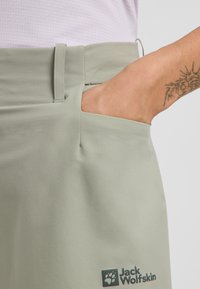 Hellgrüne Hose mit glatter Textur, ausgestattet mit einer Seitentasche und einem Logo auf der unteren linken Seite. Einfaches Design mit taillierter Taille.