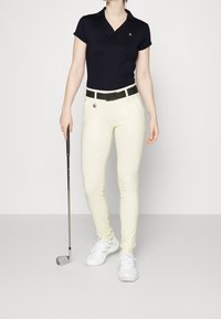 Mörkblå piké, krämfärgade byxor, svart bälte med spänne och vita golfskor. Håller en golfklubba; slankt, figurnära design genom hela.