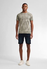 Camiseta de color verde oliva con estampado y cuello redondo, combinada con pantalones cortos de mezclilla oscura. El modelo lleva zapatillas de color claro. Diseño sencillo, tela transpirable.