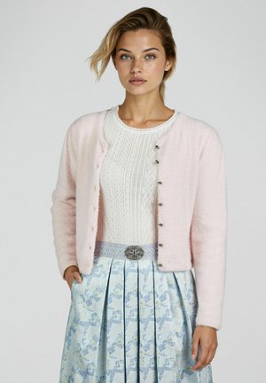 Jeune femme portant un cardigan rose clair sur un haut en dentelle blanche, associé à une jupe plissée bleue à fleurs et une ceinture décorative.