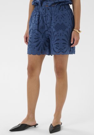 Femeie purtând pantaloni scurți albaștri brodați, pantofi mule negri cu vârf ascuțit și o brățară răsucită aurie la încheietura mâinii drepte, stând pe un fundal alb.