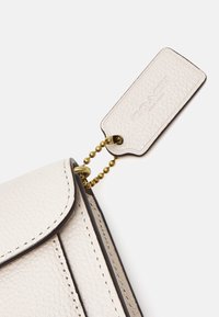 Bolso de cuero crema con un acabado texturizado, que cuenta con una cadena dorada unida a una etiqueta rectangular que muestra el nombre de la marca.