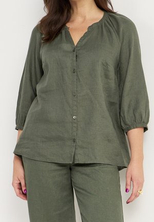 Kobieta ubrana w oliwkowozieloną bluzkę zapinaną na guziki z rękawami 3/4 z bufkami oraz dopasowane luźne spodnie, stojąca na tle jednolitej białej ściany.