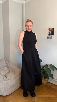 Schwarzes ärmelloses Kleid mit hohem Ausschnitt, tailliertem Oberteil und vollem Rock. Verfügt über Seitentaschen und eine glatte Textur. Getragen mit schwarzen Schuhen.