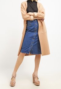 Vaalean beige pitkää kardigania mustan paidan päällä, yhdistettynä siniseen denim-lyhythelmaiseen hameeseen, jossa on etunapit ja sivusisä. Vaaleanpunaiset korkokengät.