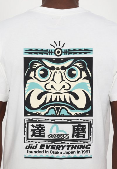 Camiseta blanca con un estampado gráfico audaz de un rostro estilizado en negro, azul y crema, con texto y elementos decorativos debajo.