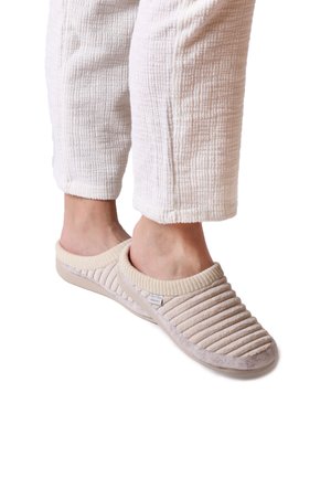 Chaussons beiges à enfiler avec une texture côtelée, un col en tricot et un matériau doux et pelucheux. Portés avec un pantalon blanc texturé.