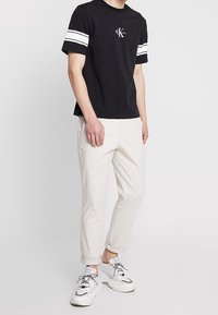 Camiseta negra de manga corta con rayas blancas en las mangas, logo de CK en el pecho. Combinada con pantalones beiges claros y zapatillas blancas.