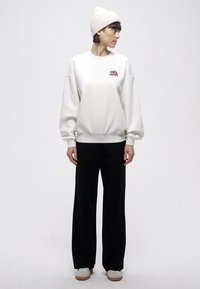 Balta sporta džemperis ar pārizmēra piegriezumu un ribveida aprocēm, ar nelielu sarkanu grafiku. Kombinēts ar melnām platām biksēm un baltiem sporta apaviem.