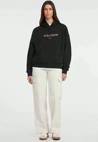 Sweatshirt preto em algodão com bolso canguru e logotipo bordado "Guess Jeans" em vermelho e branco. Combinado com calças cargo brancas e ténis.