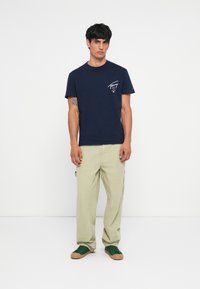 Marineblå bomulds t-shirt med et hvidt logo på brystet, parret med lysegrønne cargo-bukser og grønne sneakers. Enkel, afslappet outfit.
