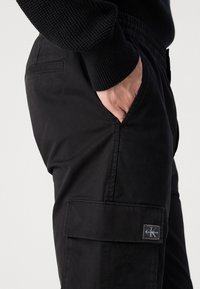 Zwarte cargobroek van gladde stof, met zijzakken, elastische tailleband en een merkpatch op de bovenbeen. Eenvoudig ontwerp.