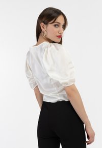 Bluză albă din satin cu mâneci umflate, guler rotund și un design ajustat. Pur tată cu pantaloni negri, expusă pe un fundal alb.