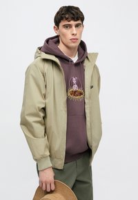 Ανοιχτό πράσινο μπουφάν με φερμουάρ πάνω από μαύρο φούτερ με γραφικό τύπωμα, συνδυασμένο με ελαιοπράσινο παντελόνι. Το άτομο κρατά ένα καπέλο σε ταμπά απόχρωση.