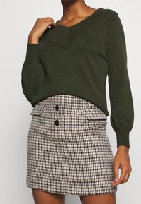 Green knitted sweater with a v-neckline, paired with a high-waisted houndstooth mini skirt featuring black buttons and side pockets.