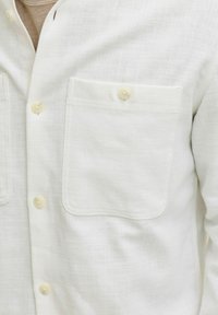 Chemise texturée de couleur claire en lin. Comprend deux poches poitrine et des fermetures à boutons avec des boutons ronds de couleur claire.